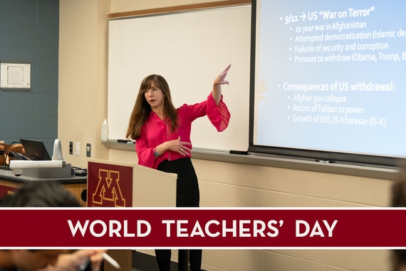 Professor dynamically pointing to a slide presentation projected at the front of a classroom. She has long brown hair and is wearing a bright pink button-down top.