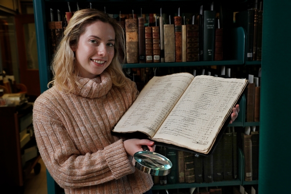 A student holds a large manuscript in one arm and a magnifying glass in the other.