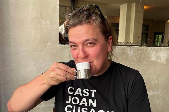 Head and shoulders of person sipping coffee; short brown hair, light skin, wearing black t-shirt with white text: Cast Joan Cusak
