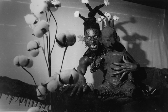 Black and white photo of a Black man dressed as a monster with batwing ears, long nails, and facepaint, holding a roughly-made child mannequin. Balls of cotton stand on stems in the foreground.