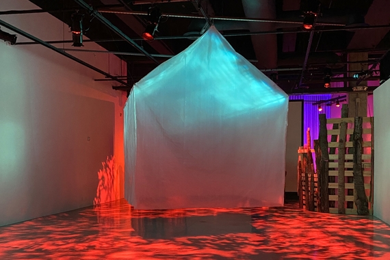 A ghostly house-shape made of plastic sheets floats in a gallery lit with red, green, and purple