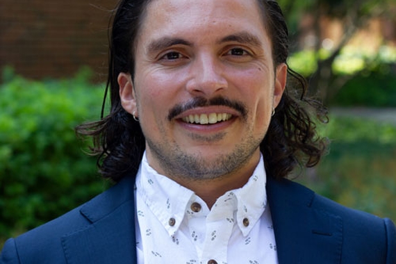 Dr. Juan Del Toro, a person with dark, shoulder length hair, and a dark mustache, wearing a blue blazer and white buttoned shirt