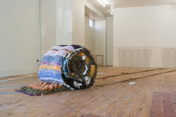 Sculptural installation of objects and hay rolled up inside a hand-woven striped rug on the floor of an unfinished room