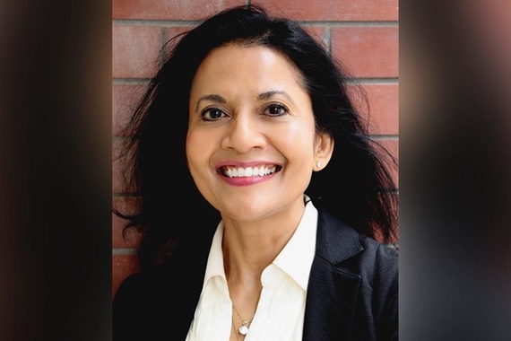 Dr. Aziz, a woman of color with black hair wearing a formal outfit, smiles in front of a brick background