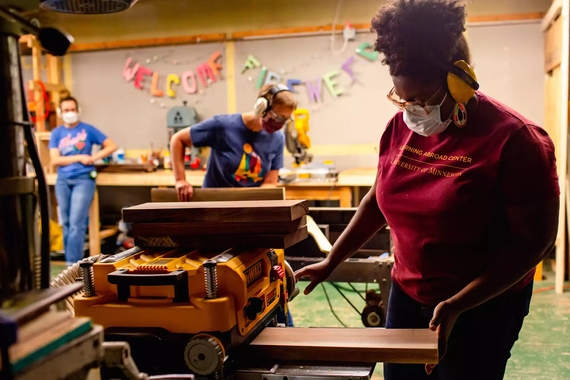 A Black woman feeds wood into a planer while a white woman uses a jointer in the background. A third looks on.