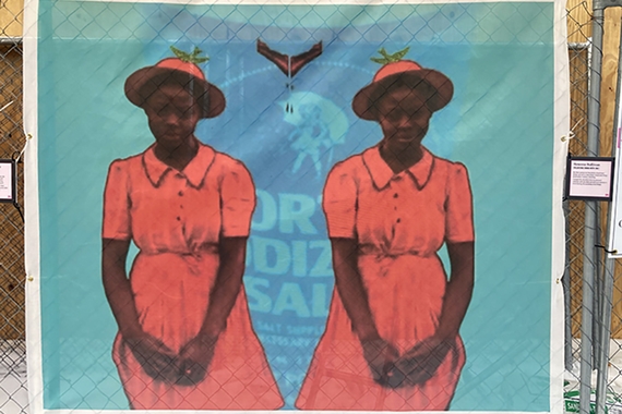Banner of two Black women in red dresses and hats with green birds on top on a blue background attached to a chain link fence