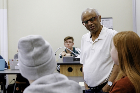 Image of classroom with two students in foreground, one in background, and focal point center right person from waist up with white hair, brown skin, wearing black glasses and white polo shirt