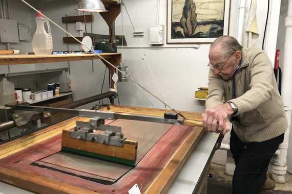 An elderly man operates the squeegee on a screenprinting press