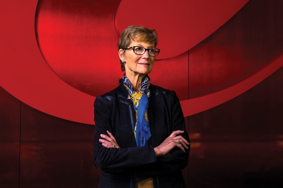 Lynn Casey wears a blue blazer and yellow scarf and stands in front of a red backdrop