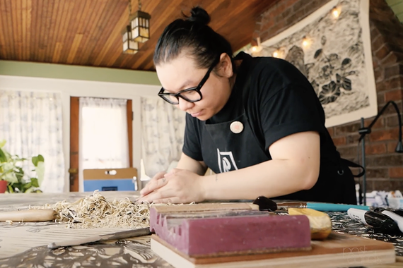 A woman in black tshirt and apron carves a woodblock