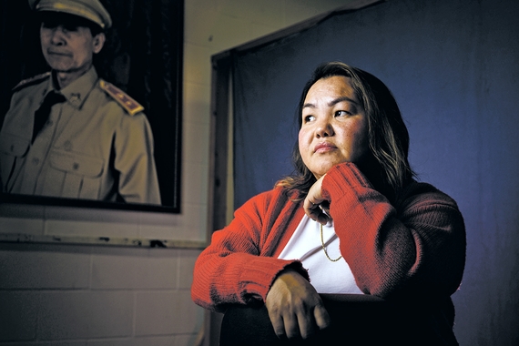 Pao Houa Her in red sweater seated in front of photo of man in military uniform