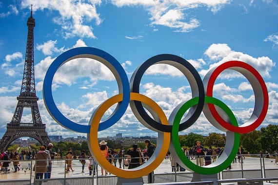 An Olympic rings sculpture with the Eiffel Towel in the background with a blue sky and clouds behind them