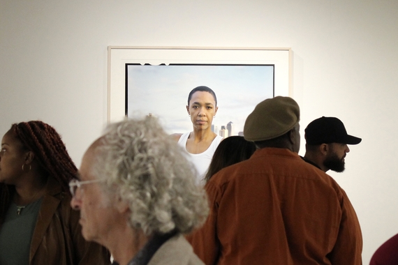 A crowd at an art gallery in front of a large photo of a gender non-conforming Black person with arms folded across their body