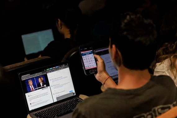 Student sitting in lecture hall with the Harris-Trump presidential debate and commentary on their laptop screen, as well as a debate bingo on their phone.