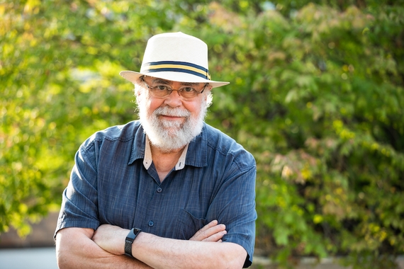 A man in a blue shirt and tan hat with glasses and a white beard looks into the camera with his arms crossed with a neutral expression on his face