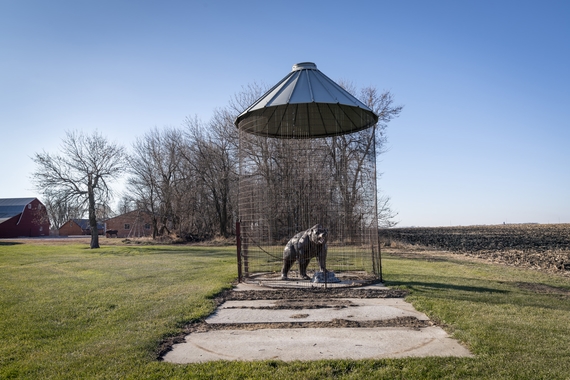 A sculpture of a bear growling stands inside a caged corn silo in a rural landscape with green grass and blue sky