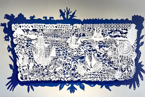 A scene of boats, waves, plants, and people cut out of white paper mounted over blue paint on a wall