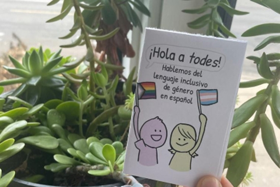 Hand holding a small zine titled ¡Hola a todos! in front of potted plants