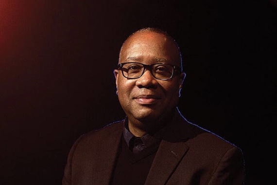 Professor Talvin Wilks smiles softly for a photo, wearing a maroon suit that blends in with the shadow-y maroon background. He wears black glasses.