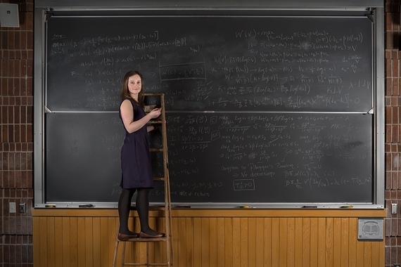 A female PhD student stands in front of a chalk board filled with equations on a tall ladder.
