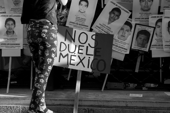 black and white image of a young woman standing before the photos of 43 students who disappeared from Ayotzinapa Mexico