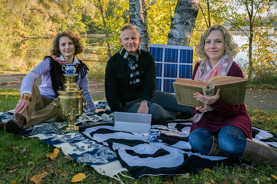 Three instructors sit in the grass at Cedar lake with a antique Russian tea set, solar cells, and a antique book.