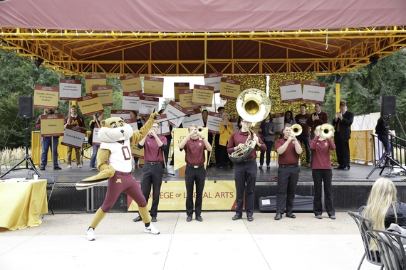 members of the marching band performing in front of a stage filled with UMN CLA faculty holding signs with their department names; Dean Coleman clapping; Goldy dancing