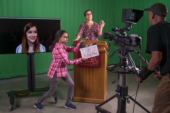 A faculty member speaks in front of a green screen while be filmed, a professional is on a video screen and a graduate student manages the room.