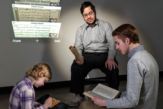 A seated graduate student instructs a child and an undergraduate with classical texts displayed on the wall.