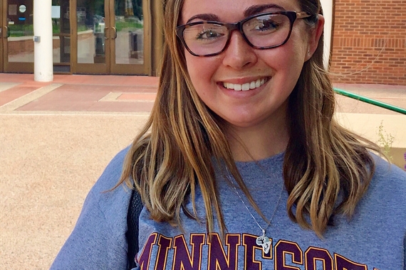 A young woman with dirty blond shoulder length hair and glasses stands in front of the doors to a brick building. She is wearing a grey t-shirt that reads "Minnesota" with a large maroon "M" outlined in gold underneath.