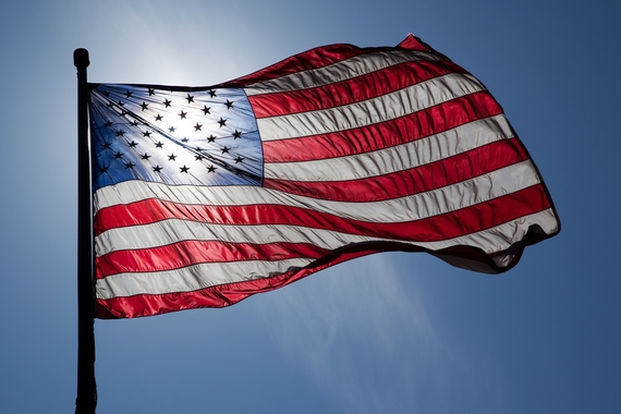 An American flag on a flagpole, blowing in the wind and blacklit by bright blue sky and sunshine