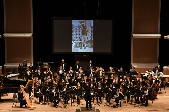 Wind Ensemble faces audience at Dr. Ben's commemorative performance. Image of Dr. Bencriscutto on projected screen above ensemble.