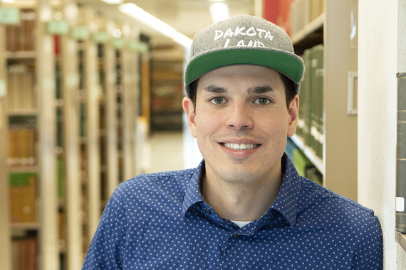 John Little smiling into the camera, standing in a library, wearing a snapback hat that says "DAKOTA LAND"