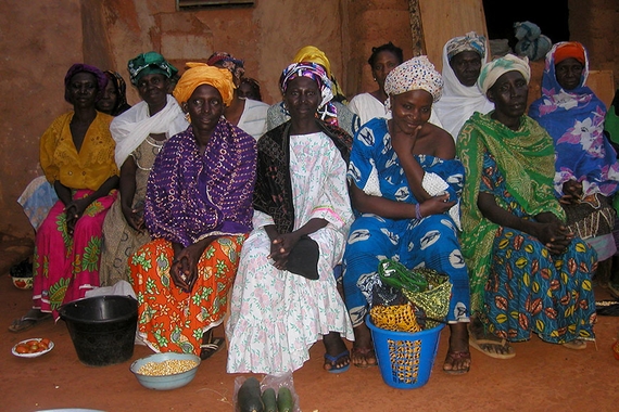 A group of Burkinese women vendors seated outside a building. They have vegetables, grains, and textiles in bowls and baskets near their feet.