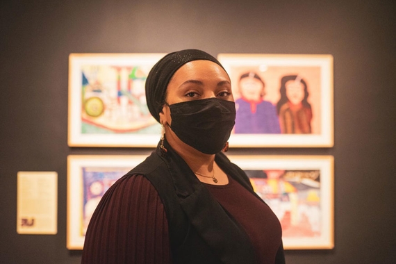 Photo of Starasea Camara. She is wearing a mask and stands in front of a group of 4 paintings.