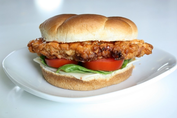 a chicken sandwich with fried chicken, tomato, lettuce, cheese, and a bun. sitting on a white plate in a white room.