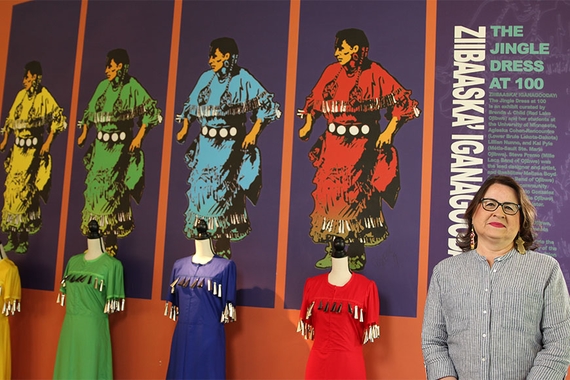 Brenda Child standing in front of a colorful display of 4 jingle dresses at a museum. Text on the display says "The Jingle Dress at 100." 
