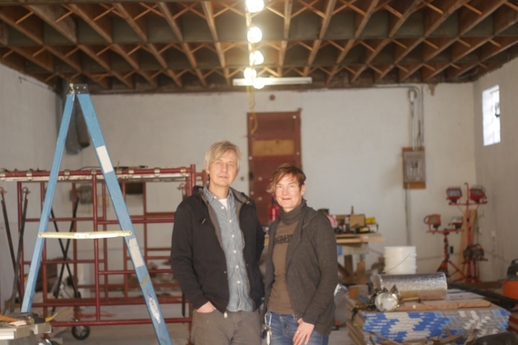 Chris Larson stands in a warehouse space next to his wife Kriss Zulkosky. There is a ladder, some lumber, and other construction materials in the space with them.
