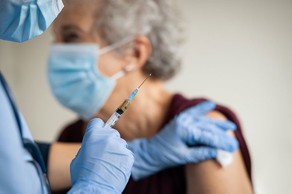 Photo of a healthcare working wearing a medical mask, gown, and gloves holding a syringe in the foreground swabbing the arm of an elderly woman in the background