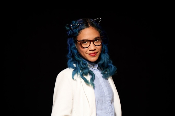 Elexis poses for camera, with blue hair, glasses, a blue shirt, and white jacket. 