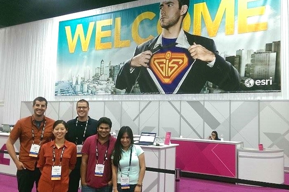 Four MGIS students at an ESRI conference. They pose in front of a large display.