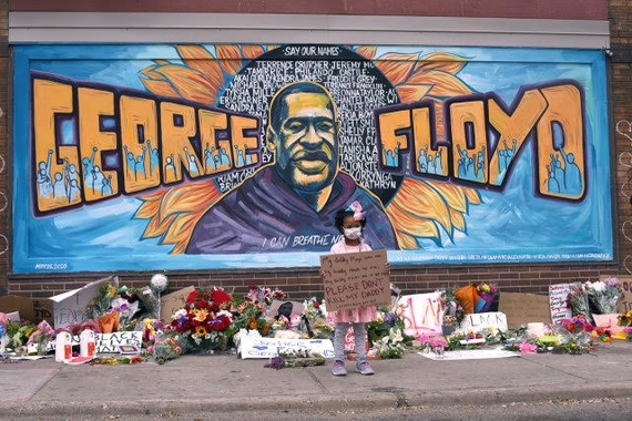 A photo of a mural depicting Minneapolis murder victim George Floyd, with a small African-American girl holding a sign in front. There are flowers and tributes on the ground in front of the mural. 