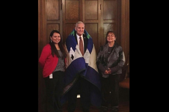 Photo of Vanessa Goodthunder standing with her mother and Minnesota Governor Mark Dayton. Dayton wears a star quilt over his shoulders