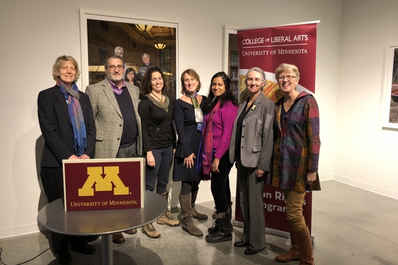 A group of seven adults stand in a line in front of a sign reading "College of Liberal Arts." There are 6 women and one man, and a podium reading "University of Minnesota" is placed on a table in the foreground.