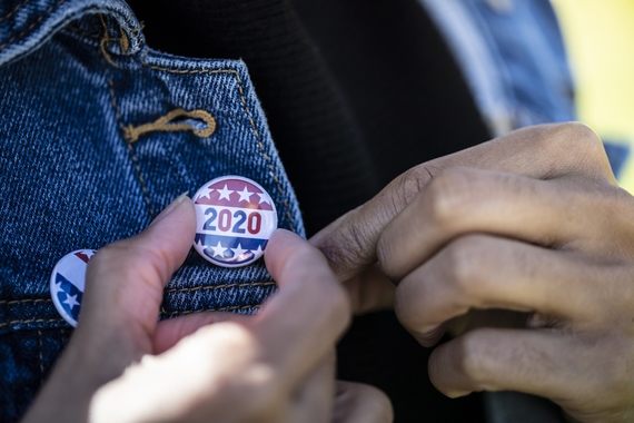 Photo of a blue jean jacket with hands putting a "Vote 2020" button on the jacket. 
