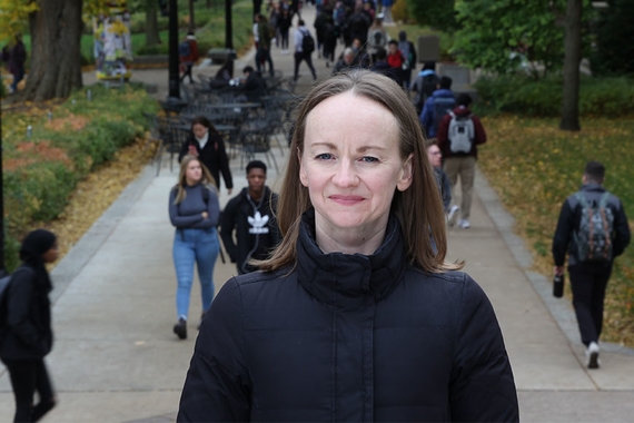 Kate Lockwood Harris looking at the camera. College students are walking on a campus sidewalk behind her.