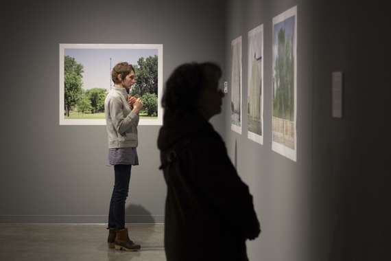 Two women look at pictures projected on the wall of an art gallery. One of trees picture can be seen behind the onlooker, but the others are somewhat obscure.
