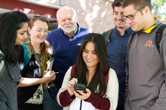 Group of people standing around a young woman who is seated. They are watching as she uses a smart phone