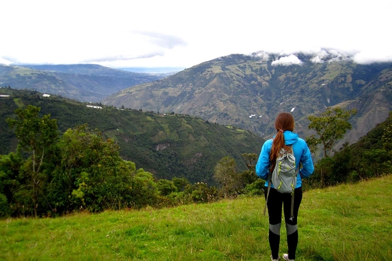 A person carrying a backpack faces away from the camera and is looking out over mountains and a valley