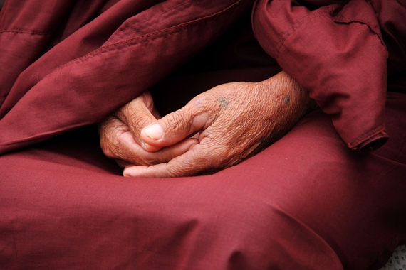 Wrinkled hands rest on top of one another in someone's lap. The person is wearing dusty red clothing, which the hands are surrounded by. A small, ilegible tattoo is seen on the left hand, at the base of the thumb. 